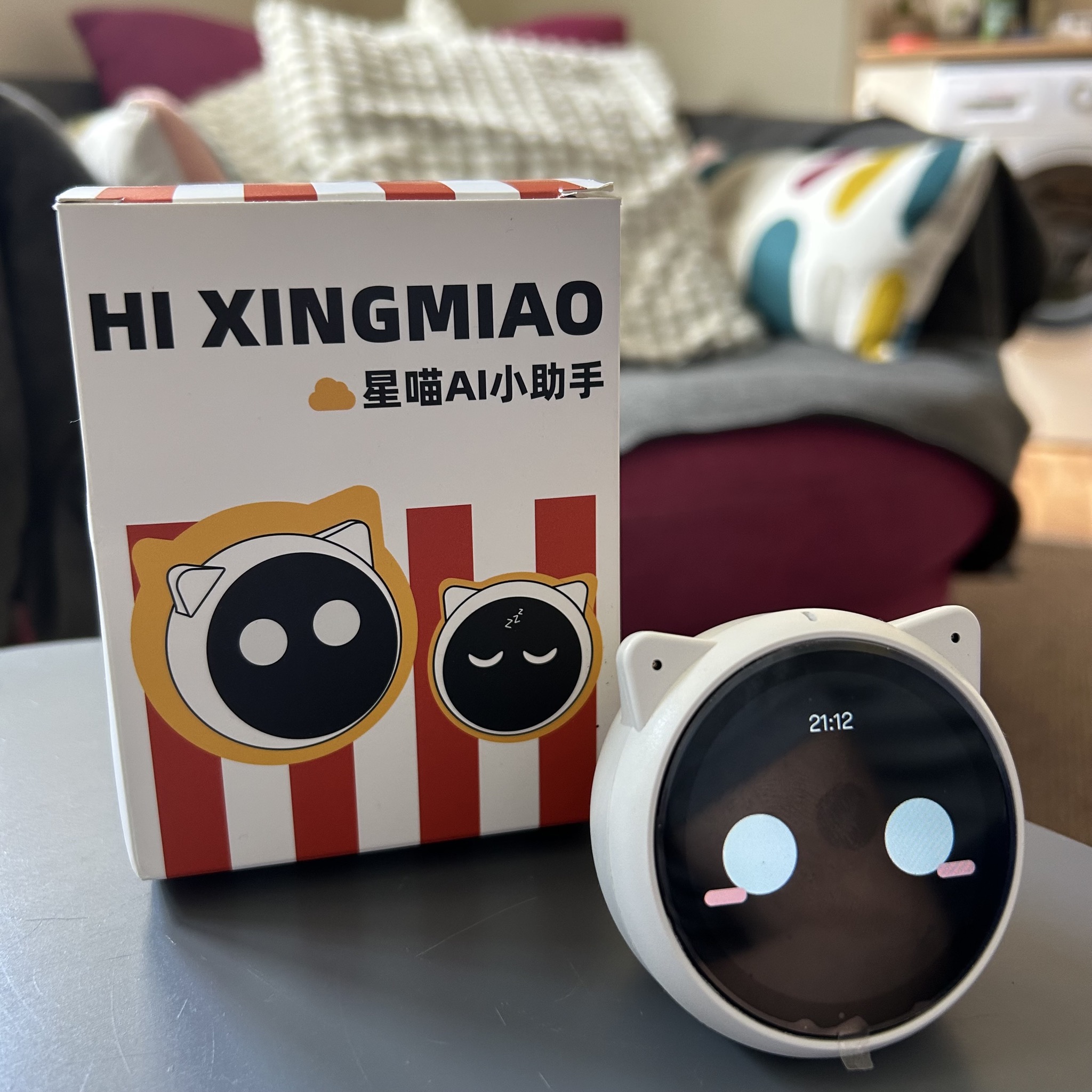 Sammy, a small round-screen speaker with white cat ears, showing pale blue eyes and pink cheeks next to its retail box labelled HI XINGMIAO